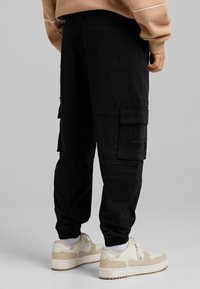 Svarta cargobyxor med flera fickor, avsmalnande anklar och elastiska muddar. Bärs med ljusa sneakers och en beige sweatshirt.