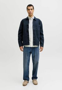 Homme debout, portant une veste navy à boutons, une chemise à rayures blanches, un jean bleu et des chaussures beige sur un fond clair uni.