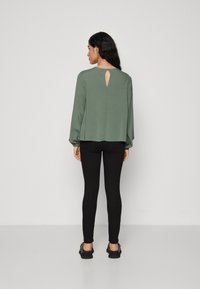 Oliven grønn langermet topp med et nøkkelhull-detalj bak, kombinert med svarte leggings og svarte sko, på en lys bakgrunn.