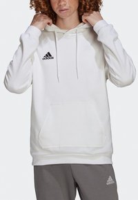 Hvit hettegenser laget av mykt stoff med snøring, med en frontlomme og en svart Adidas-logo på brystet.