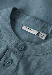 Blauwe katoenen overhemdkraag met twee blauwe knopen. Label leest "name it" en maat staat aangegeven als 1½-2J, 92. Gladde textuur en casual ontwerp.