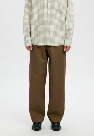 Person iført løstsiddende brune bukser, sorte sko og en langærmet beige ternet knap-til-shirt, stående mod en ensfarvet baggrund.