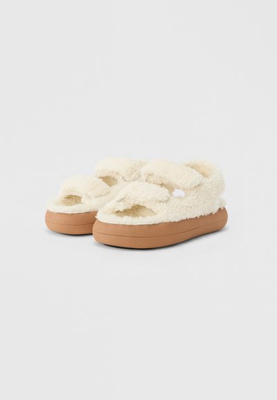 Chaussons duveteux de couleur crème avec un design à deux sangles, fabriqués en matériau doux, dotés d'une semelle en caoutchouc marron pour un confort et une adhérence accrus.