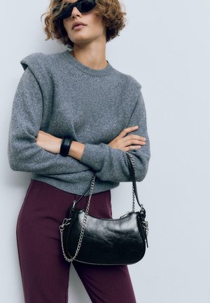 Maglione grigio con dettagli sulle spalle, abbinato a pantaloni bordeaux. Una piccola borsa in pelle nera con tracolla a catena è portata da un lato.