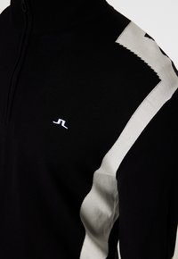 Svart zip-up tröja med hög krage, vit zigzag-design på axeln och en liten vit logotyp på bröstet. Strukturerat tyg.