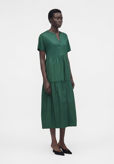 Γυναίκα όρθια, που φοράει ένα άνετο, κοντομάνικο, πράσινο φόρεμα μέχρι το γόνατο, με μαύρες μυτερές γόβες, σε απλό λευκό φόντο.