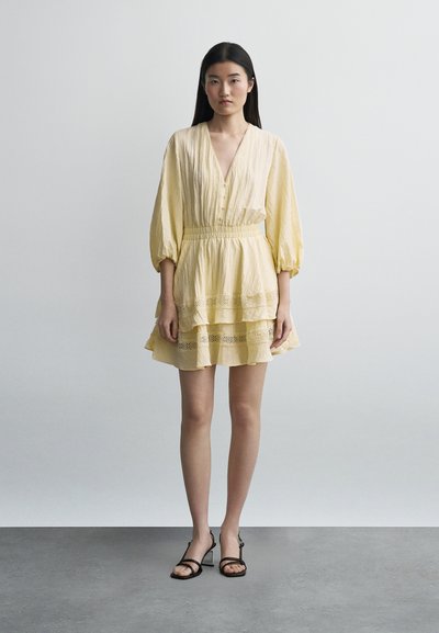 Νεαρή γυναίκα με μακριά μαύρα μαλλιά, ντυμένη με κοντό κίτρινο φόρεμα σε στρώσεις και μαύρα σανδάλια με λουράκια, στέκεται μπροστά σε απλό γκρι φόντο.