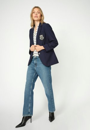 Sieviete, tērpusies tumši zilā žaketē ar ģerboni, svītrainā kreklā, zilās džinsās un melnās smailpurnu augstpapēžu zābakos, stāv uz vienkārša fona.