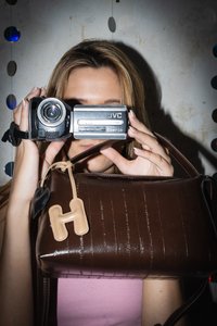 Camera video JVC Everio neagră și argintie ținută cu o mână, însoțită de o geantă dreptunghiulară maro lucioasă, cu o etichetă bej în formă de literă "H".