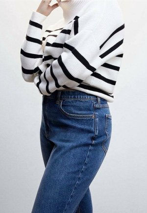 Personne portant un pull à col roulé blanc avec des rayures horizontales noires et un jean bleu taille haute, le bras plié près du visage.