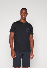 Svart T-skjorte med korte ermer laget av myk stoff, med en sirkulær lilla logo på brystet. Kombinert med mørke shorts, gir det et uformelt utseende.