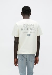 Μπλουζάκι από βαμβάκι σε κρεμ απόχρωση με γραφικό τύπωμα στην πλάτη που γράφει "DENIM ATTITUDE". Διαθέτει κοντά μανίκια και άνετη εφαρμογή.