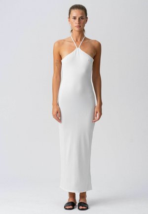 Nő áll, ujjatlan fehér halter nyakú maxiruha és fekete papucs szandál viselése közben, egyszínű világos háttér előtt.