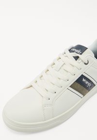 Baskets synthétiques blanches avec un design à lacets, présentant des rayures bleu marine et kaki sur les côtés et un petit logo bleu marine sur la languette.