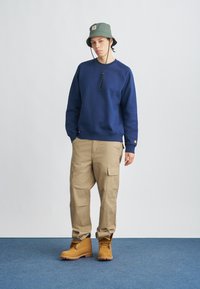 Μπλε φούτερ, μπεζ παντελόνι cargo με πλαϊνές τσέπες και κάμελ μποτάκια εργασίας. Πράσινο-ελιάς καπέλο τύπου bucket με λογότυπο. Απλός, λειτουργικός σχεδιασμός.