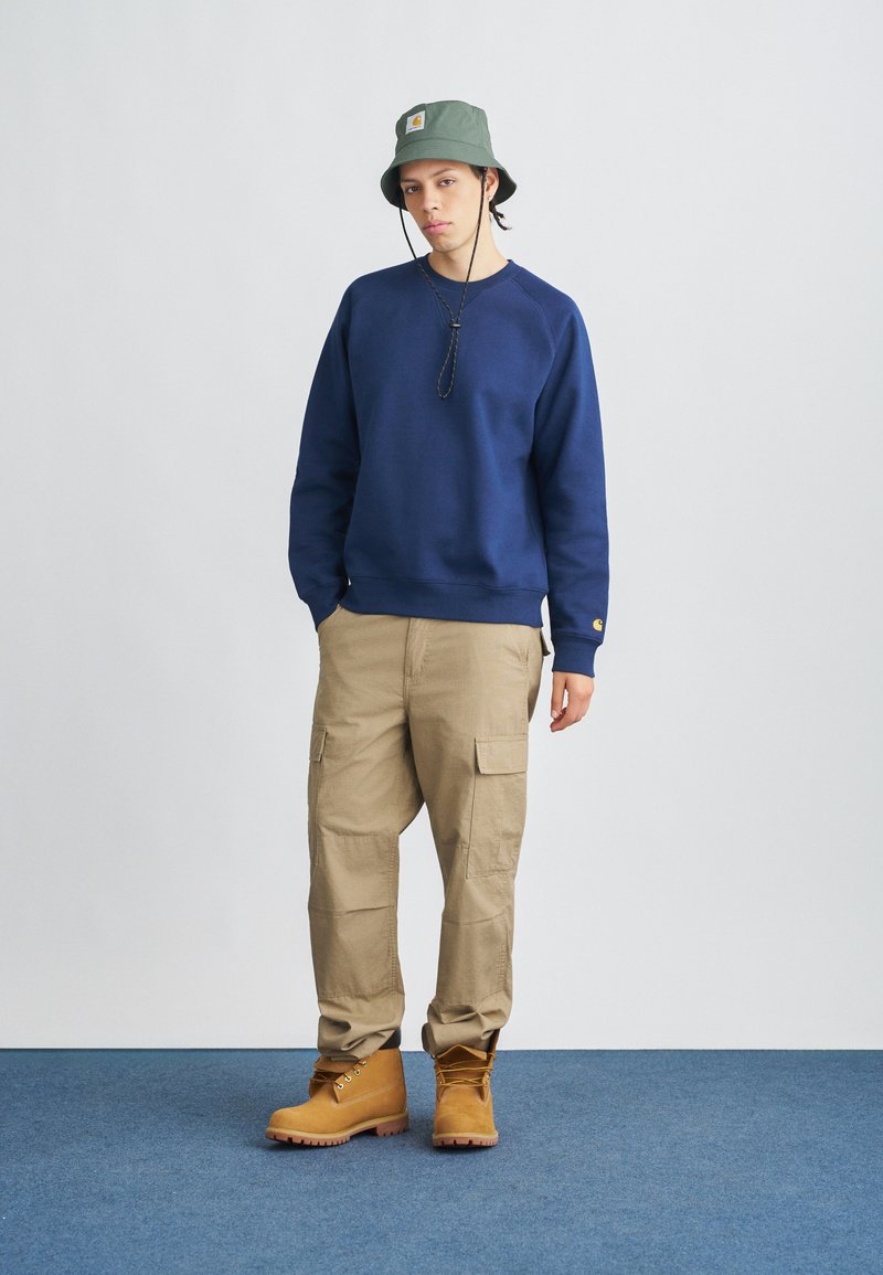 Marineblå sweatshirt, beige cargobukser med sidelommer og brune arbejdstøvler. Olivengrøn bøllehat med logopatch. Enkel, funktionel design.