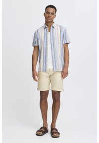 Kortearm overhemd met blauwe, roze en gele strepen; gecombineerd met beige shorts. Bruine sandalen maken de look compleet.