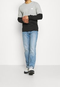 Mustapere-värillä lohkoitettu pitkähiha pullover, valmistettu puuvillasta, rento malli, ja pieni valkoinen logo-patch rinnassa. Yhdistetty vaaleansinisiin farkkuihin ja valkoisiin kenkiin.