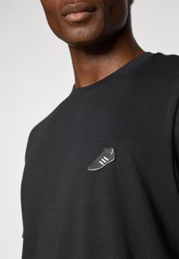 Sort t-shirt med et lille gråt og hvidt fodboldstøvlegrafik på venstre bryst. Stoffet ser blødt ud med en afslappet pasform.