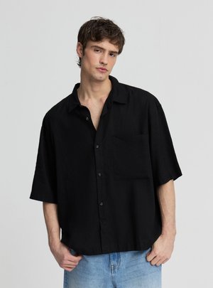 Giovane uomo con capelli castani corti che indossa una camicia nera larga a maniche corte con bottoni e jeans azzurri chiari, in piedi davanti a uno sfondo neutro.