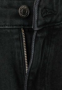 Pantalones vaqueros negros con cremallera metálica, cierre de botón y una tela sutilmente texturizada. Detalles de costura visibles a lo largo de los bordes.