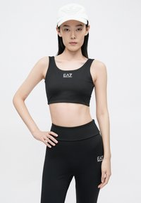 Fekete sportos crop top és magas derekú leggings EA7 logóval, rugalmas anyagból készült. A modell fehér sapkát visel dombornyomott mintával.