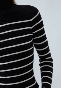 Svart ribbestrikket turtleneck-genser med smale hvite horisontale striper. Teksturen er glatt, og ermene er lange og tettsittende.