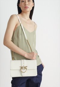 Λευκή δερμάτινη τσάντα με χρυσής απόχρωσης κυκλικό κούμπωμα, που διαθέτει αλυσίδα και καλοσχηματισμένο σχέδιο, εκτεθειμένη με μοντέλο σε χαλαρή ενδυμασία.