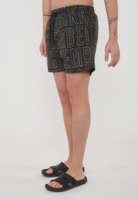 Pantaloni da bagno neri con bold testo giallo, vita elastica con cordino e fit rilassato. Abbinati a infradito nere.