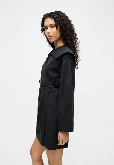 Γυναίκα με σγουρά μαλλιά που φοράει μαύρο μακρυμάνικο φόρεμα με ζώνη και φαρδύ γιακά, σε πλάγια όψη μπροστά σε απλό φόντο.