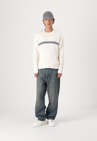 Pulover crem cu nervuri, cu dungi orizontale bleumarin, asortat cu blugi largi decolorati, de culoare albastră, și adidași albi. O șapcă gri completează look-ul.