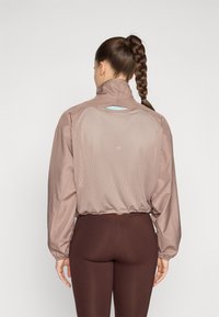 Giacca windbreaker marrone chiaro con colletto alto, schiena in rete e polsini elasticizzati, abbinata a leggins marrone scuro. Design minimalista, pratico per un uso attivo.