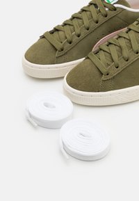 Olivově zelené semišové tenisky s texturovaným povrchem a bílými gumovými podešvemi, doplněné o dva stočené bílé tkaničky.