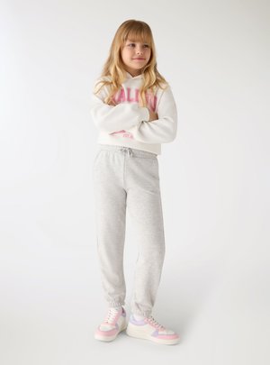 Ragazza giovane con lunghi capelli biondi che indossa una felpa bianca con cappuccio, pantaloni della tuta grigio chiaro e scarpe da ginnastica rosa pastello e viola, in piedi con le braccia incrociate.