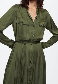 Robe vert olive, à manches longues, avec un col, deux poches poitrine, patte de boutonnage devant et une ceinture assortie à la taille. Texture de tissu douce.