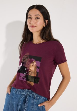 Frau mit langen dunklen Haaren trägt ein violettes Grafik-T-Shirt und blaue hoch taillierte Jeans mit Bindegürtel, steht mit den Händen in den Taschen.