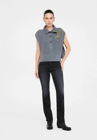 Sivý pletený sveter bez rukávov s golierom a gombíkmi, zdobený žltými kvetinovými ozdobami, v kombinácii s tmavými džínami a čiernymi topánkami.