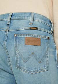 Bagsiden af lyseblå Wrangler-jeans med synligt syet "W" på lommen og en tanfarvet lædermærke med mærket på linningen mod en beige top.