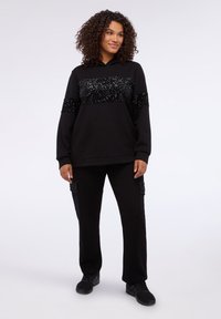 Felpa nera con dettagli in paillettes, dotata di tasca frontale e cappuccio. Abbinata a pantaloni della tuta neri con dettagli in paillettes simili.