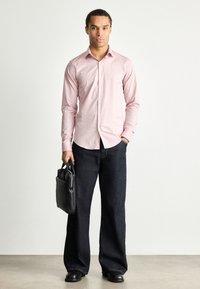 Helles rosa Langarmhemd mit Knopfverschluss, kombiniert mit dunkelblauen weitem Jeans. Eine schwarze Aktentasche und schwarze Schuhe runden das Outfit ab.