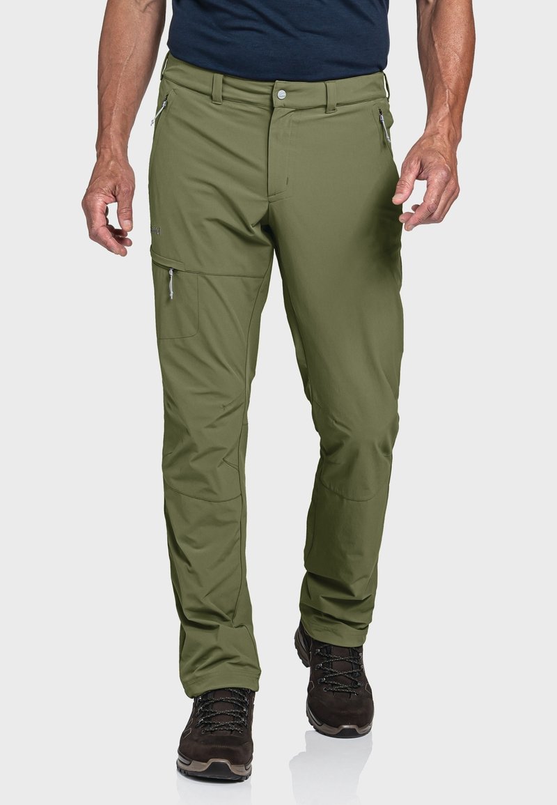 Homme portant un pantalon d'extérieur vert olive avec des poches zippées et des chaussures de randonnée noires, debout devant un fond uni.