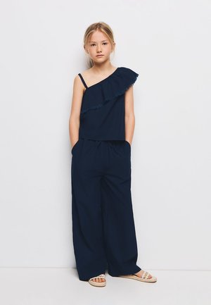 Mlada djevojka stoji sa rukama u džepovima, nosi tamnoplavi top s jednim ramenom s volanom i široke hlače, bež sandale.