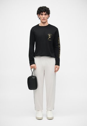 Zwart langemouwen shirt met gouden graphic, lichtgrijze broek, witte sneakers en een kleine zwarte tas. Soepele stof en een relaxte pasvorm met zijkanten.