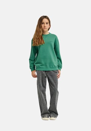 Młoda osoba stojąca w zielonej bluzie, luźnych szarych dżinsach oraz czarno-białych trampkach na jednolitym jasnym tle.