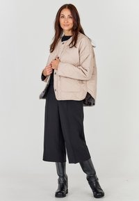 Beige quilted jacket with a round collar, front buttons, and contrast inner lining, paired with black wide-leg trousers and knee-high boots.