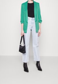 Kobieta w zielonej marynarce, czarnej bluzce, jasnoniebieskich dżinsach, czarnych botkach za kostkę i trzymająca małą czarną torebkę.