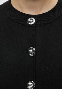 Cardigan noir en tricot avec un col rond et trois boutons argentés irréguliers brillants attachés à l'avant.