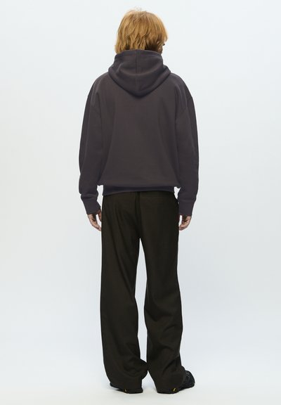 Personne aux cheveux roux portant un sweat à capuche noir, un pantalon noir ample et des chaussures noires, se tenant de dos contre un fond clair.