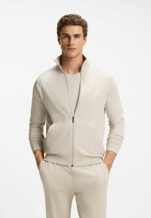 Młody mężczyzna w jasnobeżowej kurtce na zamek błyskawiczny i pasujących spodniach, z rękami w kieszeniach, stojący na jednolitym białym tle.