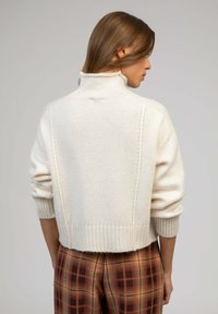 Pull en tricot de couleur crème avec un col montant, un ourlet côtelé et un motif texturé, associé à un pantalon écossais marron.